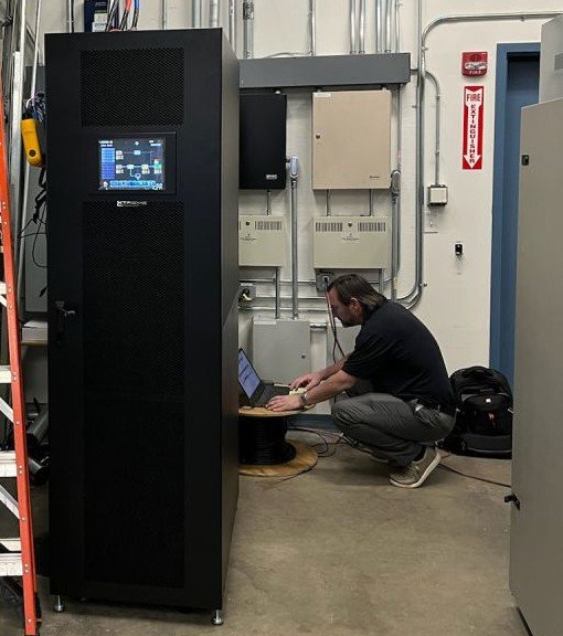 M90U-80 UPS installed in a facility electrical room, the single tall cabinet powered on with its 10-inch touchscreen showing the system one-line display, while a technician commissions the unit at a laptop alongside the building distribution panels