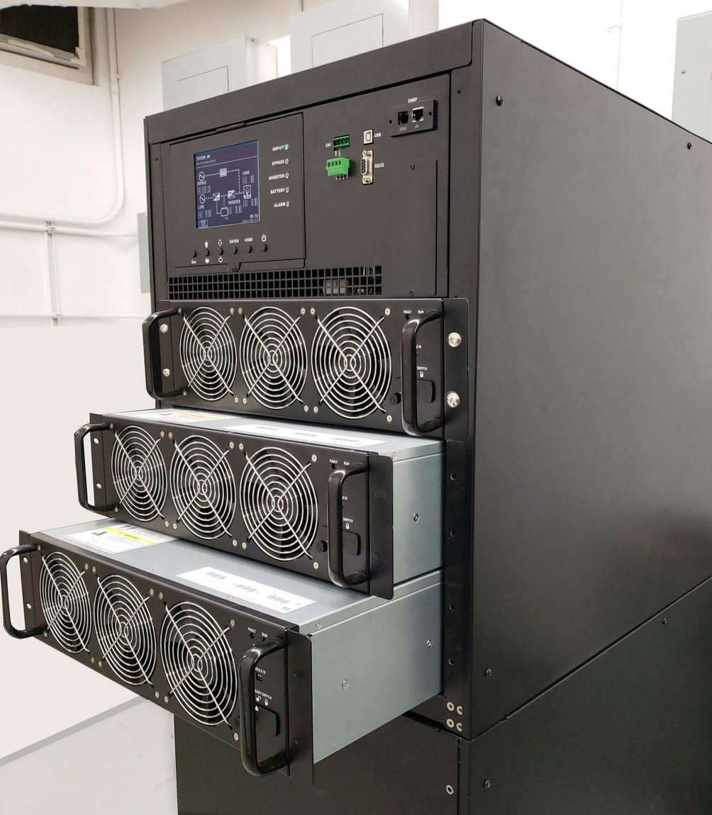 M90U UPS installed in an electrical room with its three hot-swappable power modules partially withdrawn from the enclosure, each module showing its three-fan array and pull handles, above the integrated control panel with graphic LCD and SNMP/USB/RS-232 communications