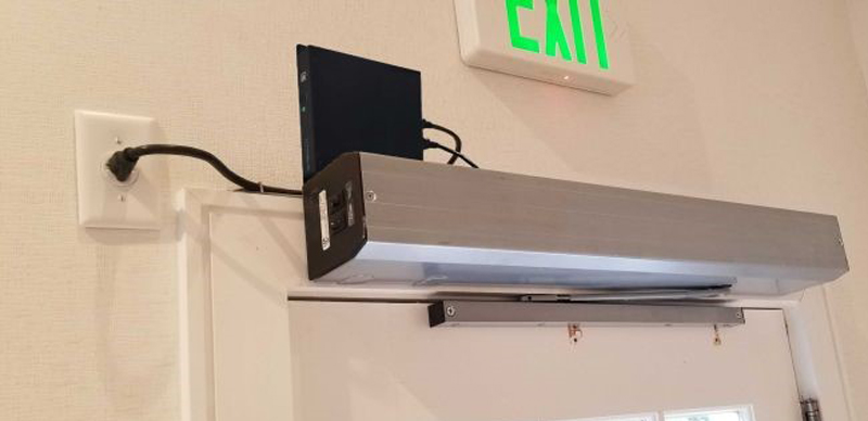 J60 lithium UPS mounted on wall directly above automatic door operator — exit sign above, door frame and closer visible below, real installation