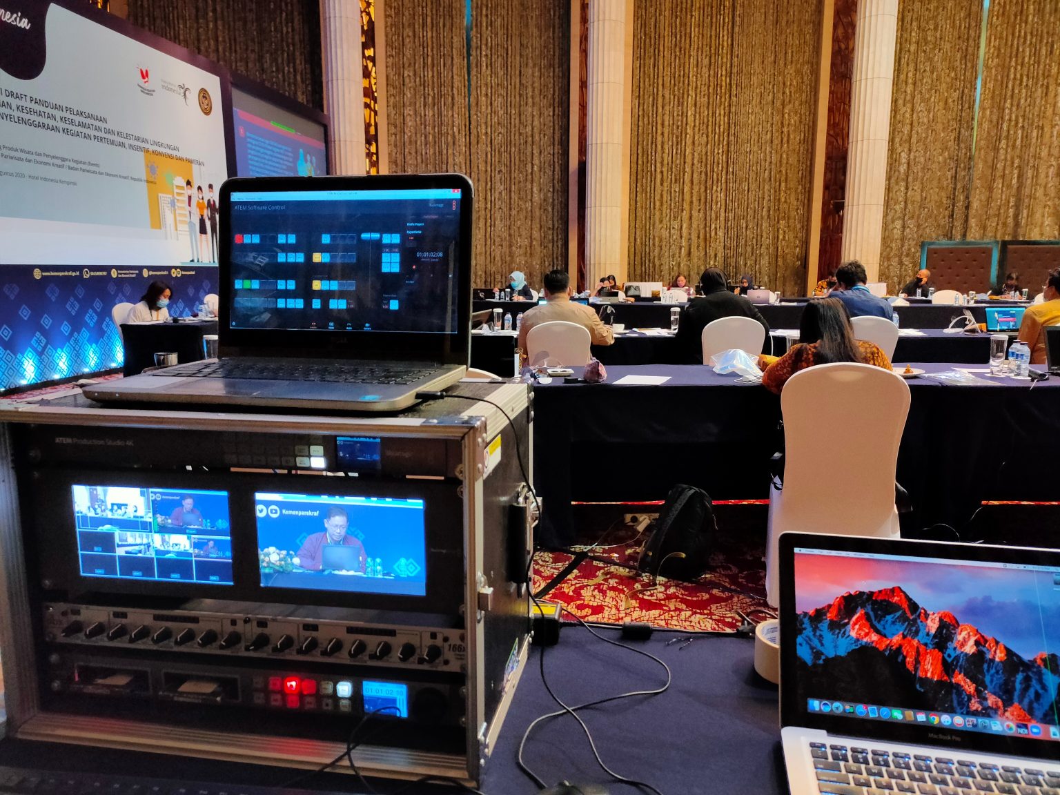 Hotel ballroom conference AV production setup — ATEM Production Studio 4K in flight case rack with live video monitoring in foreground, conference delegates and projection screen in ballroom background