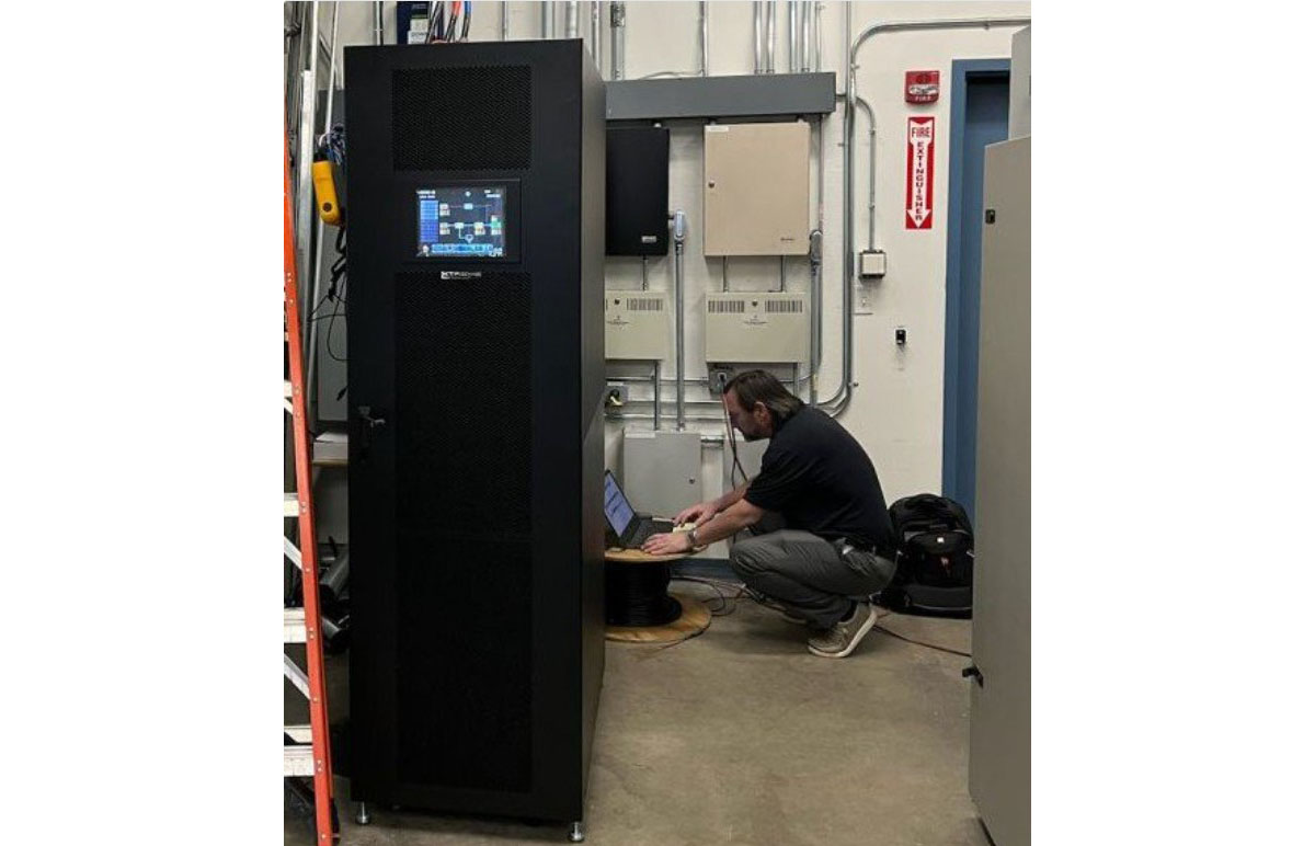 M90U-80 UPS installed in a facility electrical room, the single tall cabinet powered on with its 10-inch touchscreen showing the system one-line display, while a technician commissions the unit at a laptop alongside the building distribution panels